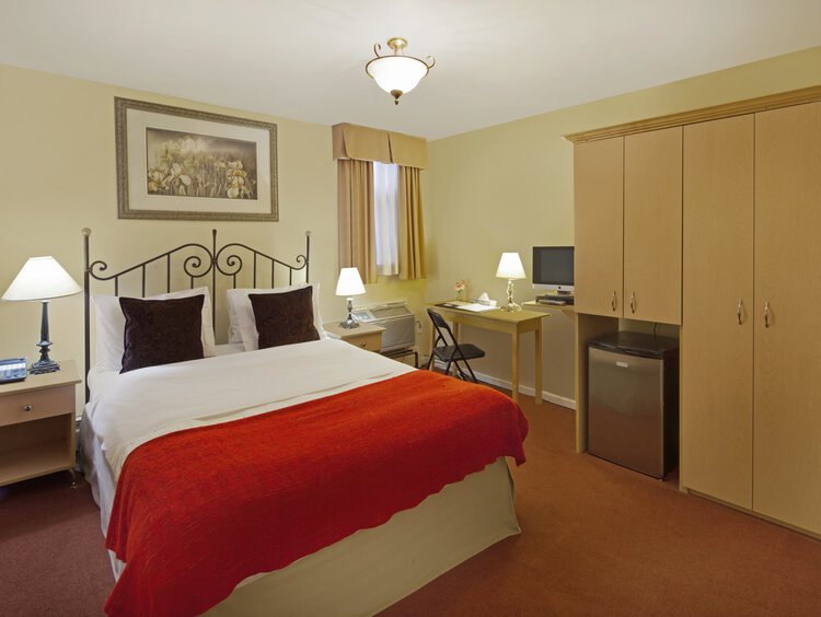A guest room with a queen bed, nightstands, a desk, and mini-fridge