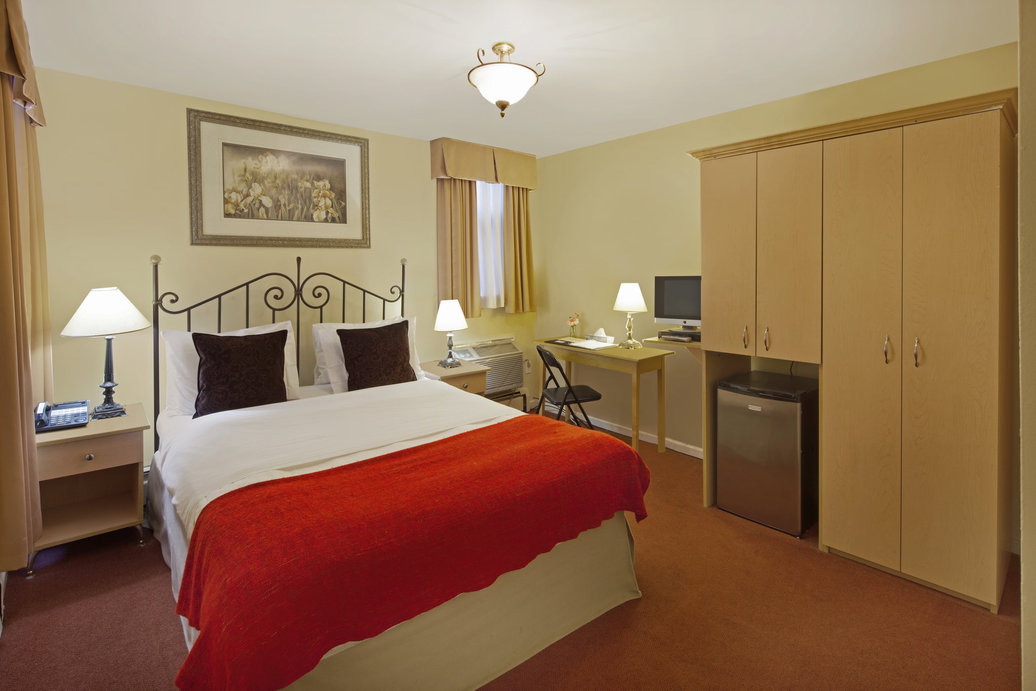 A guest room with a queen bed, nightstands, a desk, and mini-fridge